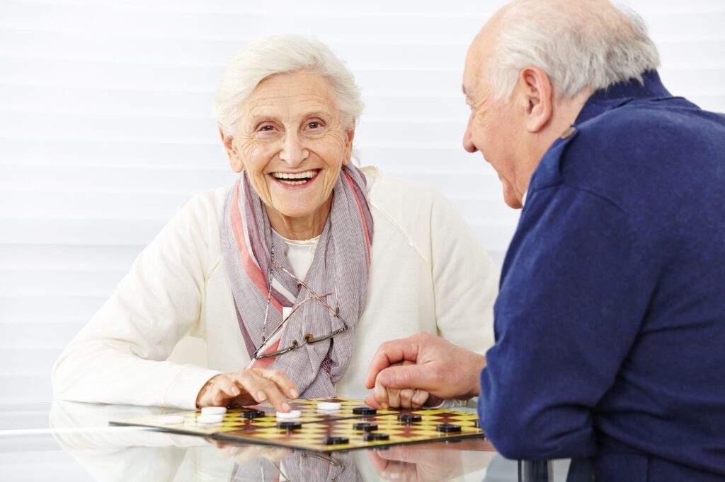 Two smiling older adults sitting at a table playing a board game called checkers.