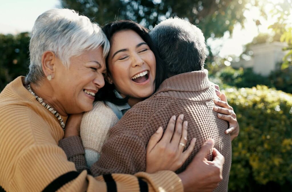 An adult daughter hugs her senior parents outside of their home in an assisted living community.