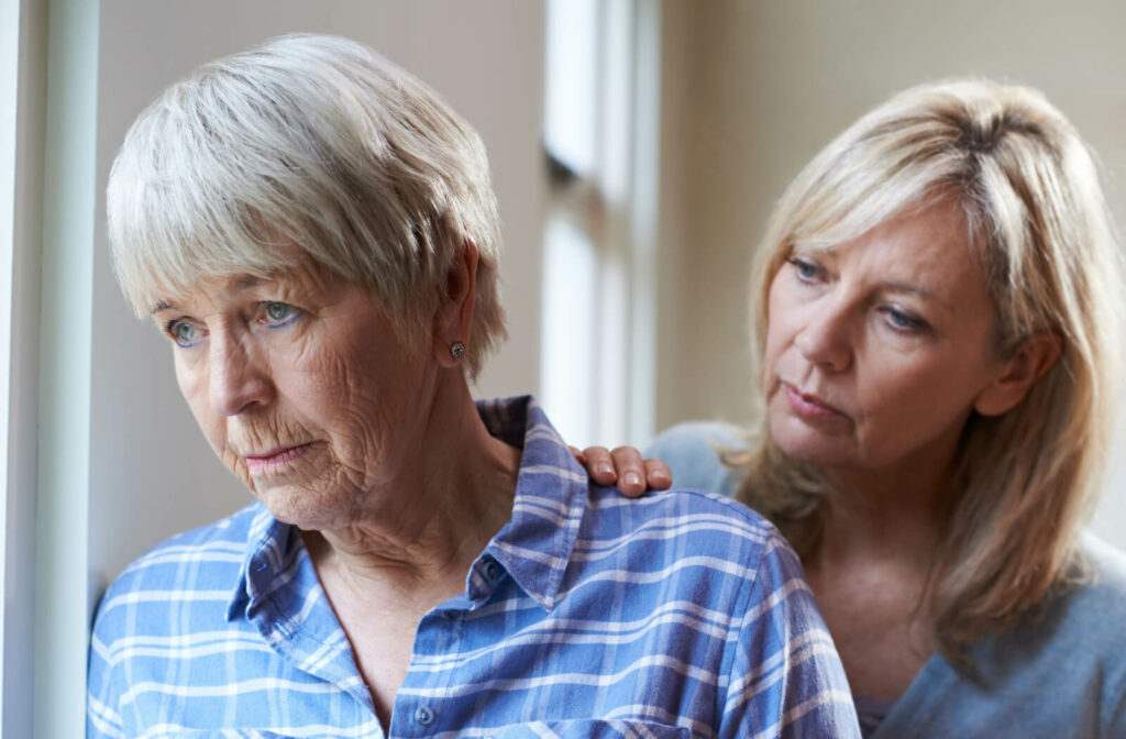 An older adult with dementia staring out the window in frustration while their adult child rests a hand on their shoulder to support them.